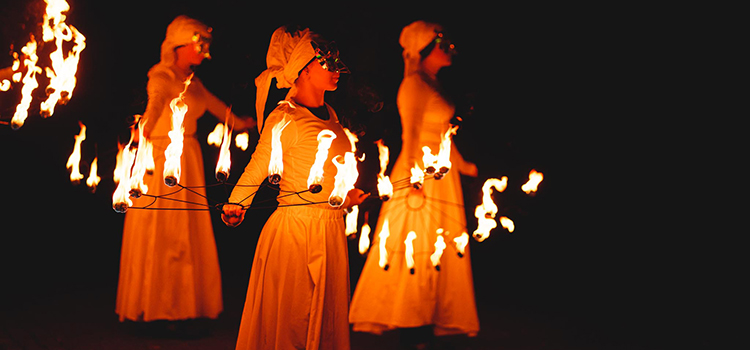 En grupp scenklädda personer med smyckade ansiktsmasker håller i föremål som brinner