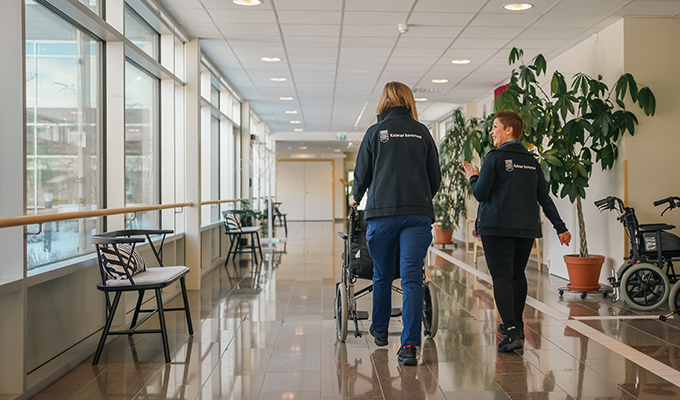 Två personer går bortåt i en ljus korridor i ett vård- och omsorgsboende och pratar med varandra. De har på sig arbetskläder med Kalmar kommuns logotyp. En av dem kör en rullstol.
