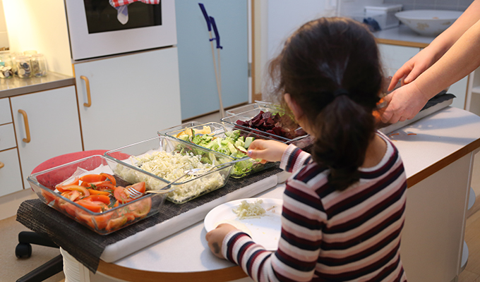 Ett barn i randig tröja med ryggen vänd mot kameran tar för sig av grönsaker. Platsen ser ut att vara ett förskolekök.