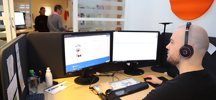 En man  med headset sitter i förgrunden framför en datorskärm. I bakgrunden syns en kvinna ta emot besök i receptionsdelen av kontaktcenter.