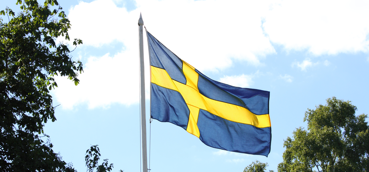Närbild på svenska flaggan mot ljusblå himmel i bakgrunden. 