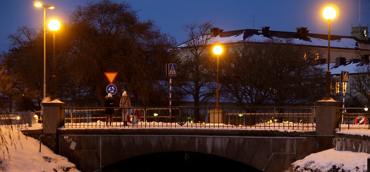Två personer i mössa står på bron vid stationen i Kalmar. Det är kväll och gatulamporna lyser.